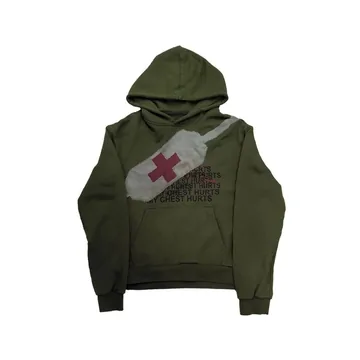 An olive green hoodie hanging on a white wall, with a red cross print on a pale panel across the chest.